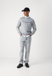 Pelēks džemperis ar "TOMMY HILFIGER" logotipu, zem tā balta krekla apkakle, gaiši pelēkas bikses un melni apavi, papildināti ar melnu cepuri.