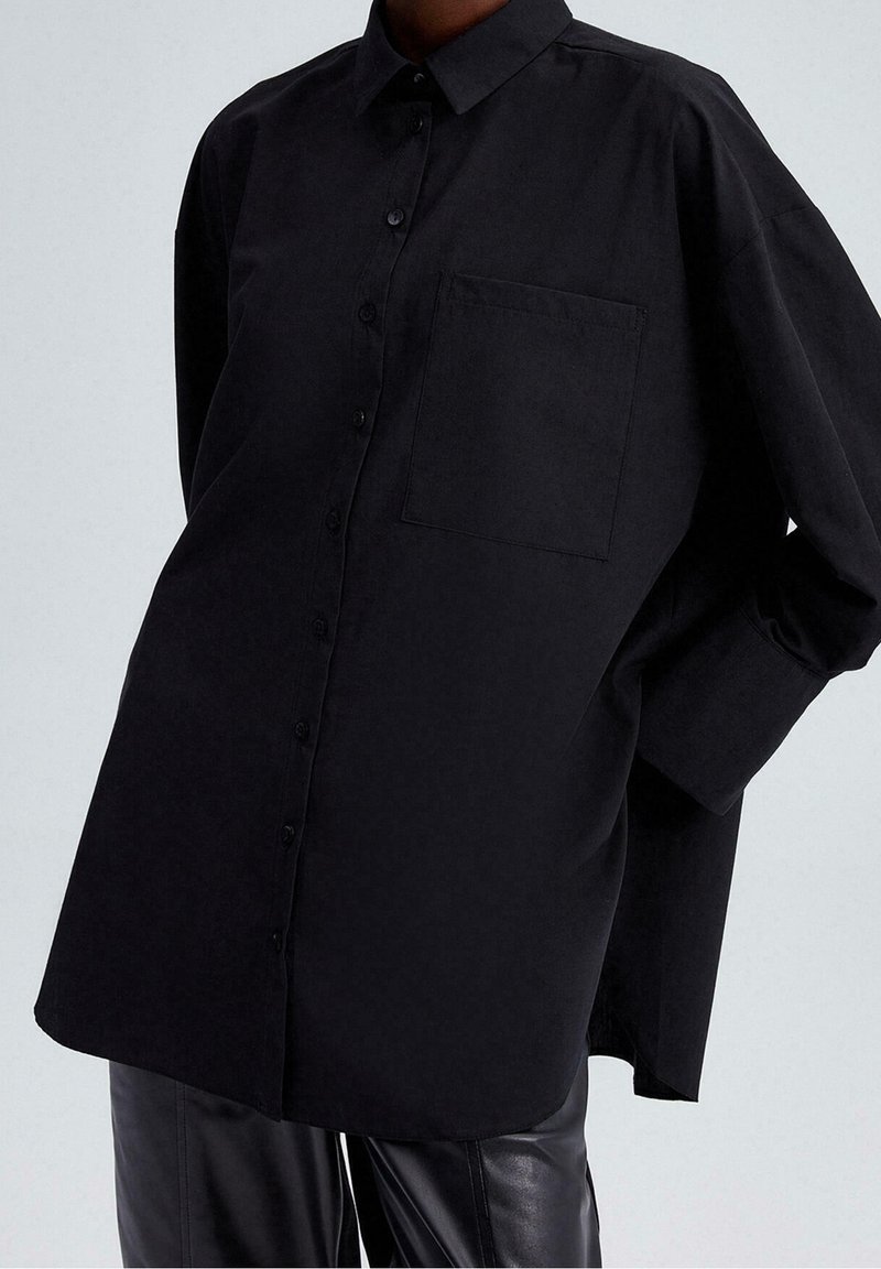Personne portant une chemise noire oversize à boutons avec une poche poitrine et un pantalon en cuir noir sur un fond clair uni.