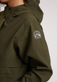 Olivgrön vattentät jacka med huva. Har en slät yta, knäppning med tryckknapp och en cirkulär logotyp på vänster ärm med bergdesign.