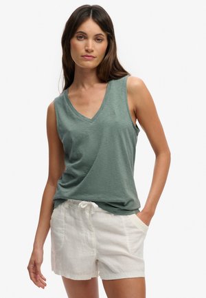 Vrouw draagt een mouwloze groene top met V-hals en witte shorts met trekkoord, staand met één hand in haar zak tegen een effen achtergrond.