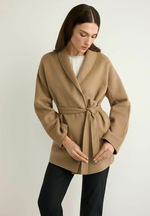 Kobieta w camelowym, wełnianym płaszczu z paskiem, nałożonym na biały top i czarne spodnie, stojąca na jasnojednolitym tle.