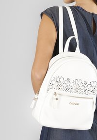 Valkoinen keinonahkainen reppu, jossa on kukkaelokuvaihanteet, kultaiset yksityiskohdat, kaksi vetoketjullista osiota ja säädettävät valkoiset hihnat.