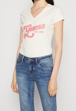 Frau trägt ein weißes V-Ausschnitt T-Shirt mit pinkem "Guess EST. 1981" Schriftzug und blaue Jeans, steht vor einem schlichten Hintergrund.