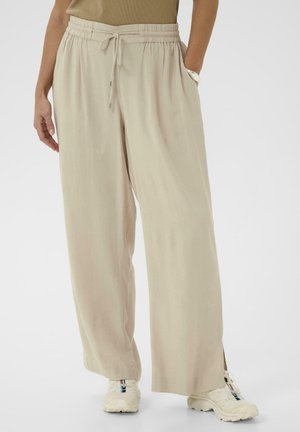 Pantalon beige à jambes larges avec cordon de serrage, porté avec des baskets blanches et un haut marron, une main dans la poche et un bracelet argenté visible.