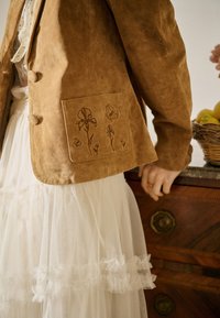 Femeie purtând o jachetă maro din piele întoarsă cu broderie florală pe buzunar și o fustă albă din tul stratificat, stând lângă o comodă din lemn.