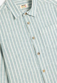 Camicia a righe blu chiaro e bianca con bottoni in legno, dotata di colletto e taschino sul petto. Realizzata in tessuto intrecciato.