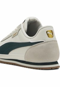 Hvide sneakers med mørkegrønne akcenter, ruskindshæl og struktureret overdel. Har guld-logo og gummisål med et karakteristisk mønster.