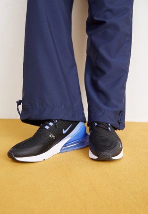 Persoon die zwarte en blauwe Nike Air Max 270 sneakers draagt met marineblauwe broek, staand op een geel tapijt.