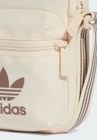 Borsa in tela beige con logo Adidas marrone, scompartimenti con zip e tracolla a righe. Presenta un tessuto testurizzato e dettagli di branding sottili.