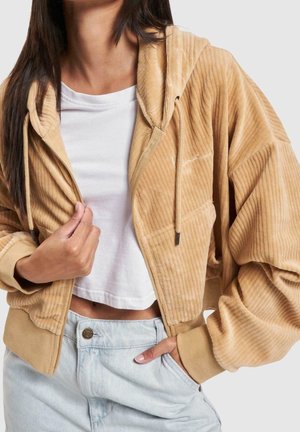Vrouw draagt een beige corduroy rits hoodie over een wit crop top en lichtblauwe spijkerbroek met hand in zak tegen een effen achtergrond.