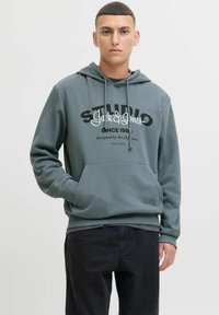 Hoodie vert en tissu doux, avec une poche avant, des cordons de serrage et un texte graphique blanc en gras. Associé à un pantalon sombre.