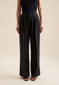 Czarne szerokie spodnie z lekkiego materiału, z wysokim stanem i plisowanym przodem, zestawione z czarnymi sandałami.