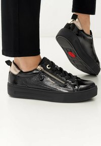 Fekete lakkbőr sportcipők cipzár részletekkel és arany díszítéssel. Vastag fekete talp; a saroknál található kis húzófül megkönnyíti a felvételt.
