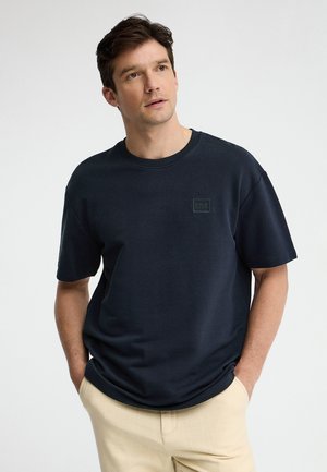 Mann trägt dunkelblaues Polo Club T-Shirt und beige Hosen, Hände in den Taschen, schaut leicht nach oben und zur Seite.