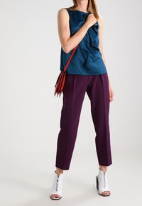 Turkusowa bluzka z falbanami, fioletowe dopasowane spodnie i białe botki za kostkę z zamkiem. Czerwona torebka crossbody podkreśla stylizację.