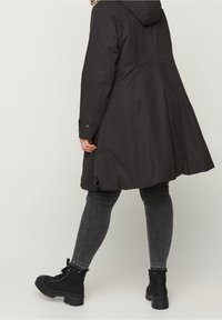 Cappotto impermeabile nero con cappuccio, design svasato e maniche con bottoni. Abbinato a jeans scuri e stivaletti neri con lacci.