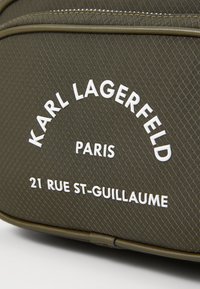 Zaino in tessuto verde con texture, dal design arrotondato, caratterizzato da testo bianco in rilievo: "KARL LAGERFELD PARIS 21 RUE ST-GUILLAUME."
