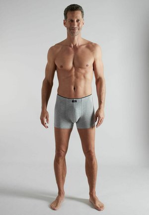 Fit middle-aged man standing barefoot, wearing gray boxer briefs, smiling against plain light gray background.