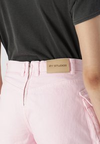 Jeans din denim roz deschis cu o etichetă din piele bej pe care este înscris "2Y STUDIOS." Detalii includ un fermoar, bucle pentru curea și buzunare plate.