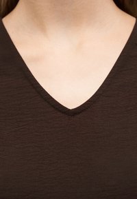 Κοντινό πλάνο του λαιμού και του πάνω μέρους του στήθους ενός ατόμου που φοράει σκούρο καφέ μπλουζάκι με V λαιμόκοψη και εμφανή υφή υφάσματος.