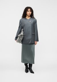 Γκρι σακάκι προσαρμοσμένης κοπής με μοναδικό ασύμμετρο σχέδιο, φορεμένο πάνω από πράσινη σατέν φούστα και συνδυασμένο με μαύρες μπότες μέχρι το γόνατο. Κρατάει τσάντα από δέρμα φιδιού.