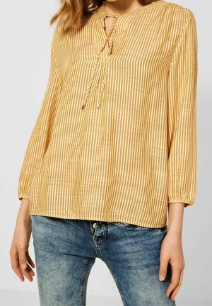 Kvinde iført en gul og hvid vertikalt striped bluse med lange ærmer og V-hals med bindebånd foran, kombineret med blå falmede jeans.