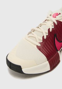 White athletic shoe featuring a textured cream upper, dark red accents, black sole, and pink detailing on the side. Laced design with mesh texture.