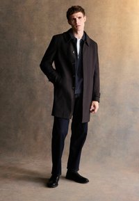 Brązowy płaszcz wełniany z kołnierzem, zapinany na guziki, z bocznymi kieszeniami, noszony na granatowym swetrze i ciemnych dżinsach z czarnymi butami.