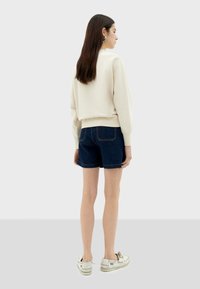 Pull crème avec ourlet côtelé, associé à un short en denim foncé avec des coutures visibles et des revers. Chaussures blanches à lacets.