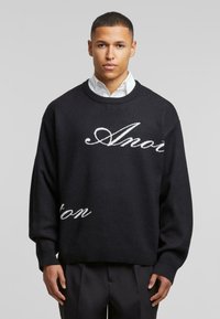 Suéter negro de punto con letras en cursiva blancas en la parte delantera. Presenta un cuello redondo y puños y dobladillo acanalados.