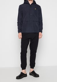 Personne portant une veste à capuche noire zippée, un pantalon de jogging noir et des sabots noirs à enfiler avec des clous métalliques autour des semelles.