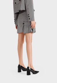 Zapatos de tacón alto de charol negro con punta afilada y tacón grueso. El diseño presenta un acabado brillante y una silueta elegante.