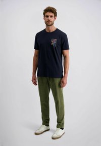Man met kort krullend haar staat in een zwart graphic t-shirt, groene corduroy broek en witte sneakers tegen een effen witte achtergrond.