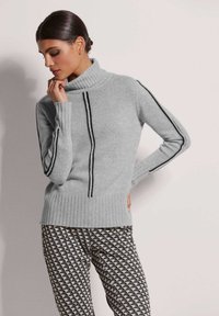 Grauer gestrickter Rollkragenpullover mit schwarzen vertikalen Streifen, gerippten Bündchen und Saum, kombiniert mit geometrisch gemusterten Hosen in Schwarz und Weiß.