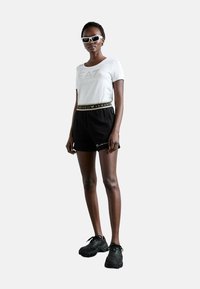 T-shirt bianco a maniche corte con logo stampato, pantaloni corti neri con cintura in evidenza dorata e scarpe da ginnastica nere. Gli occhiali da sole bianchi completano il look.