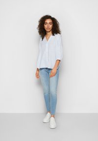 Camisa de botões azul claro com riscas, de corte descontraído e mangas de três quartos, combinada com jeans skinny de lavagem clara e ténis brancos.