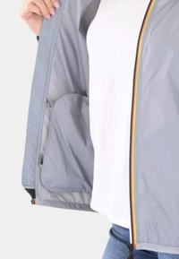 Chaqueta ligera gris con cremallera de rayas multicolor, detalles de bolsillo interior y textura suave. Llevada sobre una camisa blanca y jeans azules.