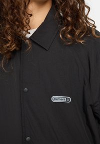 Zwarte lichtgewicht jas met een kraag, drukknoopsluiting en een zichtbaar "Element" logo patch aan de zijkant. Gladde textuur, slank ontwerp.