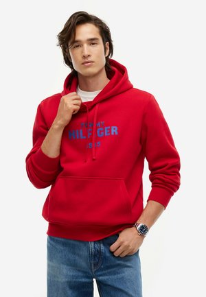 Giovane uomo con felpa rossa Tommy Hilfiger e jeans blu con orologio da polso, posato su sfondo bianco semplice.