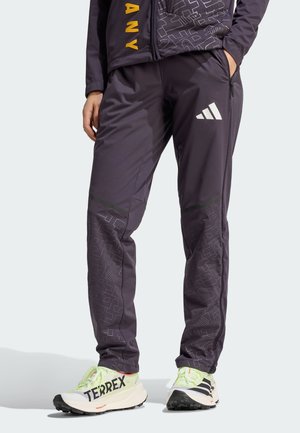 Person trägt schwarze Adidas-Trainingshose mit weißem Logo und gemusterten Details, kombiniert mit hellen Adidas Terrex Sneakern.