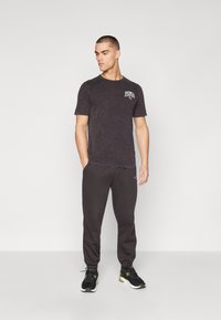 Schwarzes T-Shirt mit einem verblassten Muster und weißen Puma-Logo, kombiniert mit schwarzen Jogginghosen und schwarzen Sportschuhen mit grünen Akzenten.