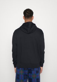 Hombre de espaldas vistiendo un sudadera con capucha negra y pantalones de cuadros azules contra un fondo blanco liso.