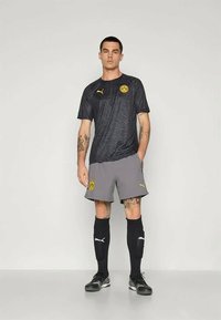 Fekete sportmez finom mintával, sárga logóval. Szürke rövidnadrág, fekete zokni és fekete focicipő. Karcsú fazon, sportos stílus.