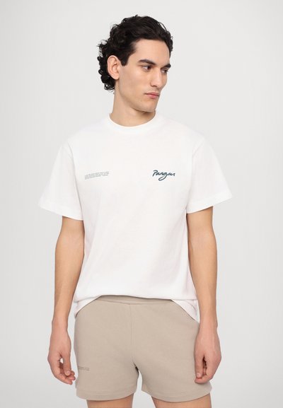 Weißes Baumwoll-T-Shirt mit Rundhalsausschnitt, ausgestattet mit einem türkisfarbenen Logo und Textdesign auf der linken Brustseite, kombiniert mit beigen Shorts.