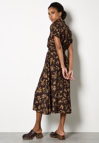 Abito midi marrone a fiori con design a maniche corte e bottoni, vita elastica e tessuto testurizzato; abbinato a zoccoli marroni.