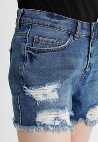 Modré džínové šortky s třepenými lemy a poškozenými místy, nošené s černým topem, ukazující detaily bočních a předních kapes.