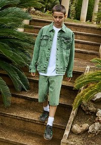Giacca di jeans verde e pantaloni corti abbinati indossati su una maglietta bianca. Scarpe nere con accenti verdi e calzini bianchi. Sfondo di gradini di pietra e vegetazione.