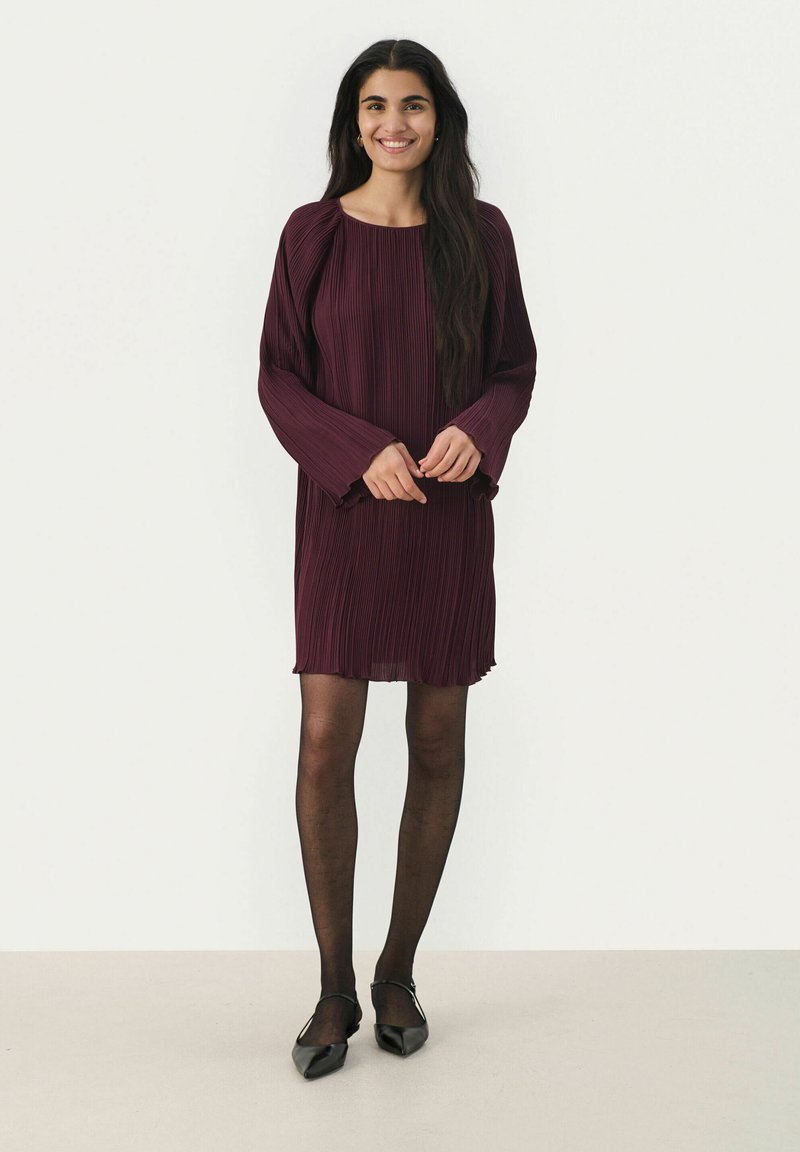 Burgunderfarbene Plisseekleid mit langen Ärmeln, rundem Ausschnitt und einem Saum, der über das Knie fällt. Getragen mit schwarzen flachen Schuhen und transparenten schwarzen Strumpfhosen.