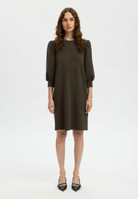 Tummanvihreä, pehmeästä kankaasta valmistettu hihallinen mekko, polvipituinen suoralla leikkauksella ja pyöreällä kaula-aukolla, yhdistetty mustiin teräväkärkisiin kenkiin.