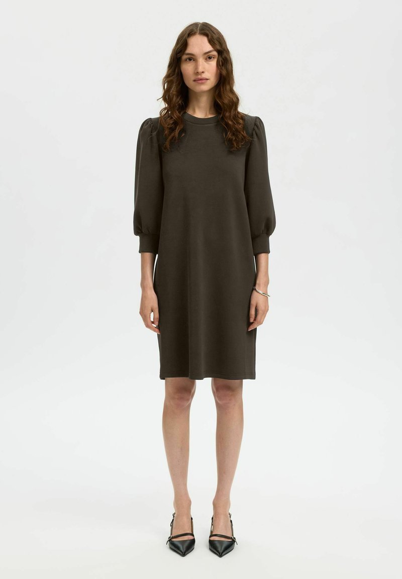 Tummanvihreä, pehmeästä kankaasta valmistettu hihallinen mekko, polvipituinen suoralla leikkauksella ja pyöreällä kaula-aukolla, yhdistetty mustiin teräväkärkisiin kenkiin.